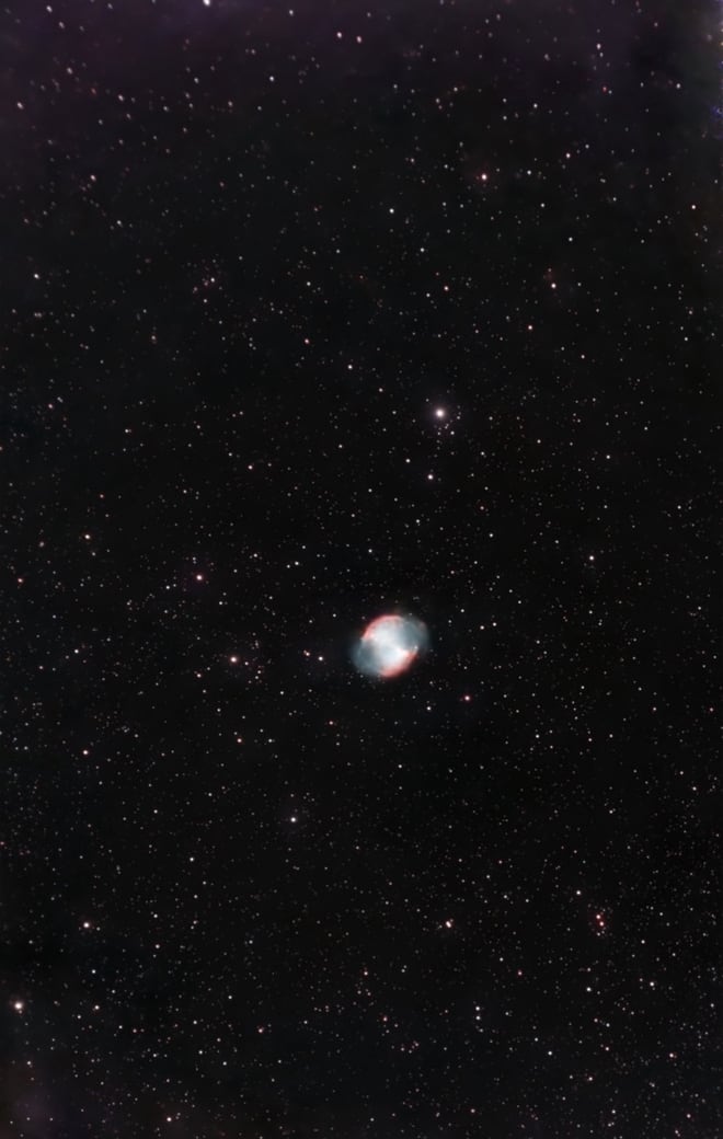 The Dumbbell Nebula: a small, bright blue and pink planetary nebula shaped like an apple core, alone in a dense starfield.