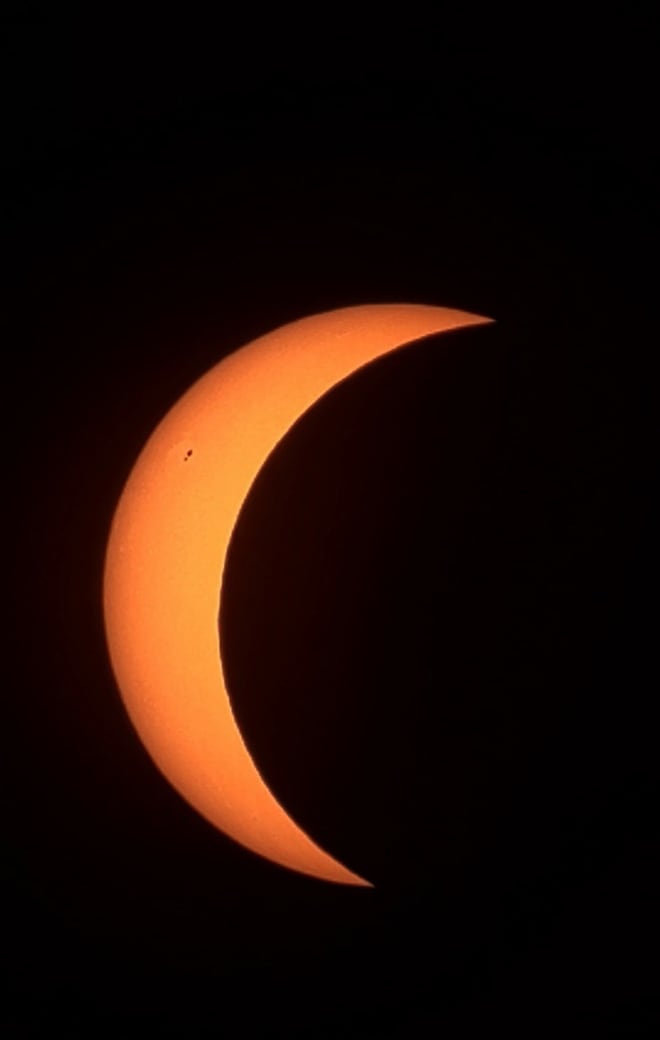 The Sun as a thin orange crescent, most of its disc hidden by the Moon, with a small sunspot near the limb.
